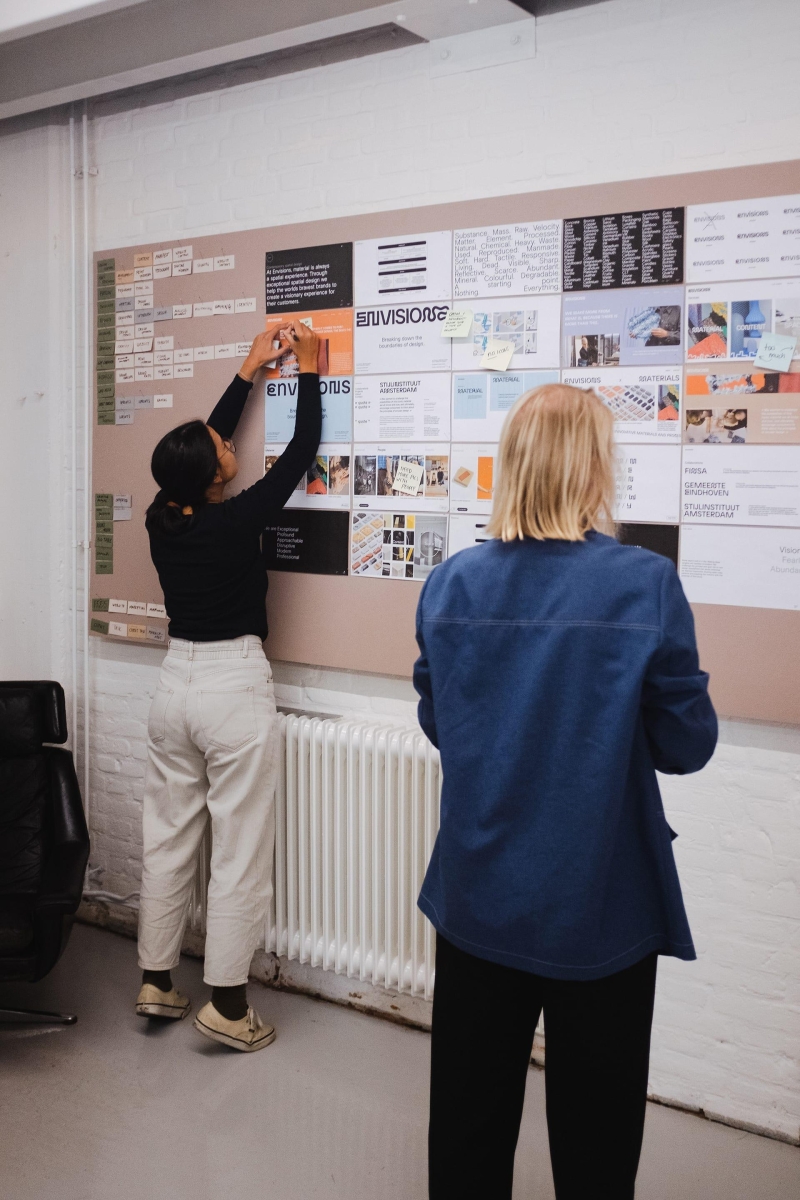 Two graphic designers sharing their work on a new branding project on a pinboard in a creative design studio.